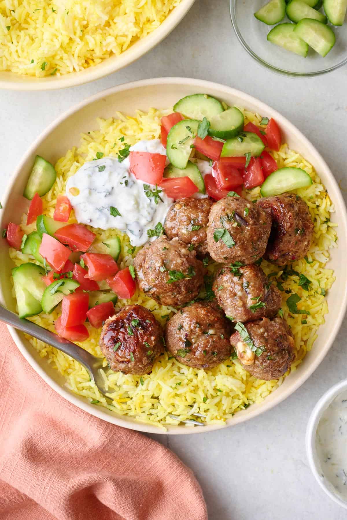 Serving of lamb meatballs with feta on a plate over Lemon Rice with mint tzatziki sauce spooned on top, chopped roma tomatoes and cucumbers on side.