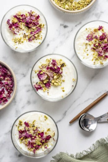 Four servings of muhallabia, or milk pudding, in small glasses garnished with crushed pistachios and rose petals.