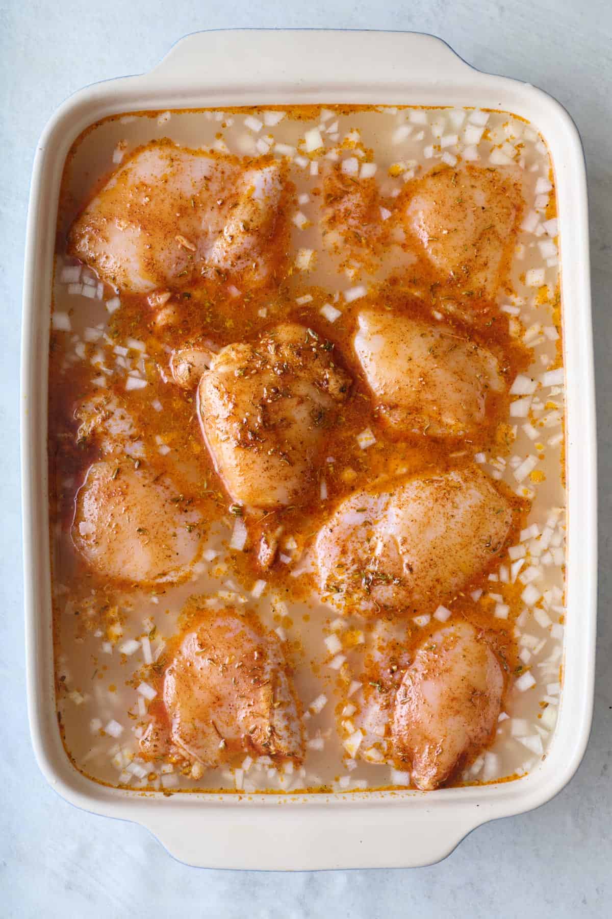 Seasoned chicken thighs added on top of uncooked rice in water in baking dish.