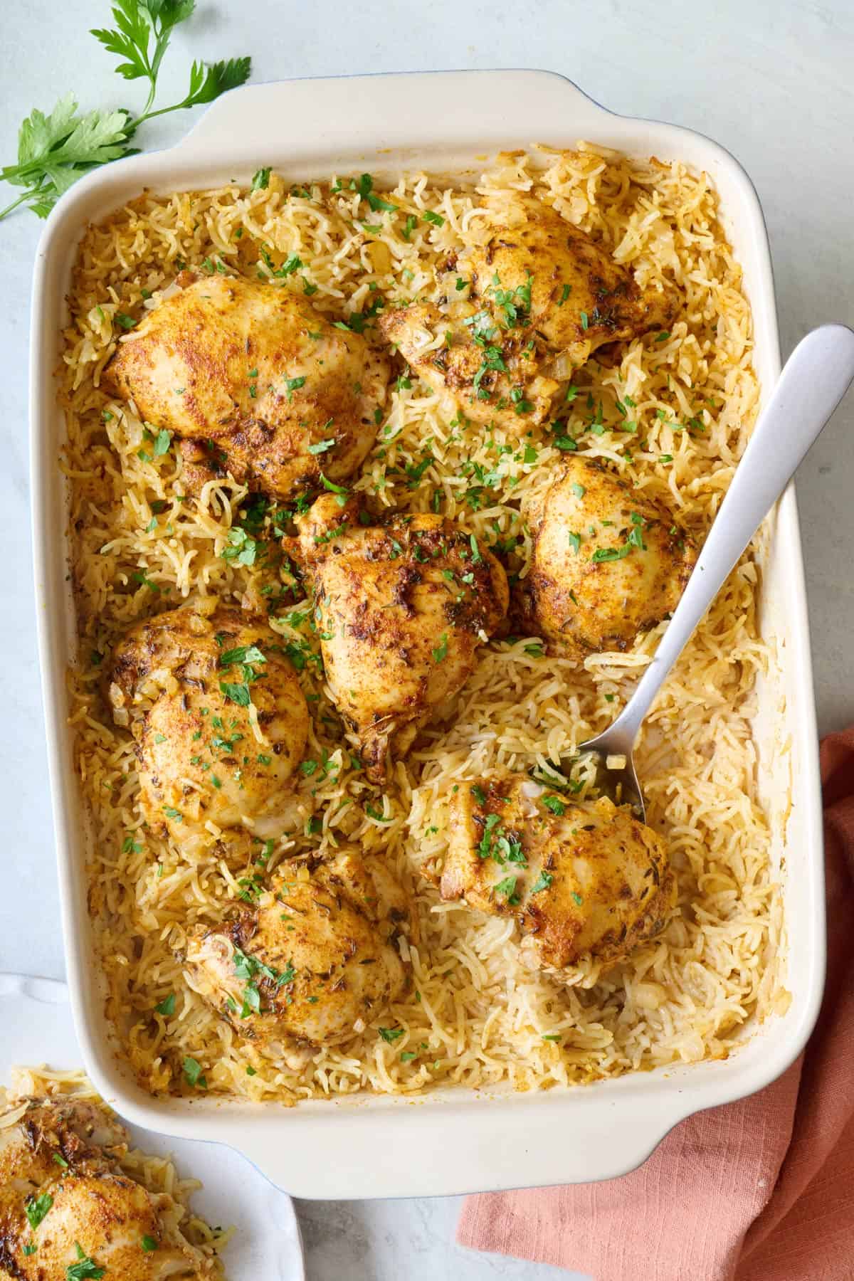 Serving spoon dipped into baking dish of chicken and rice with serving of recipe on a plate nearby.