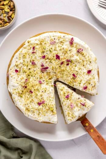 Homemade pistachio cake topped with cream cheese frosting, crushed pistachios and rose petals with a slice cut and pulled away from cake, small dish of pistachios nearby.