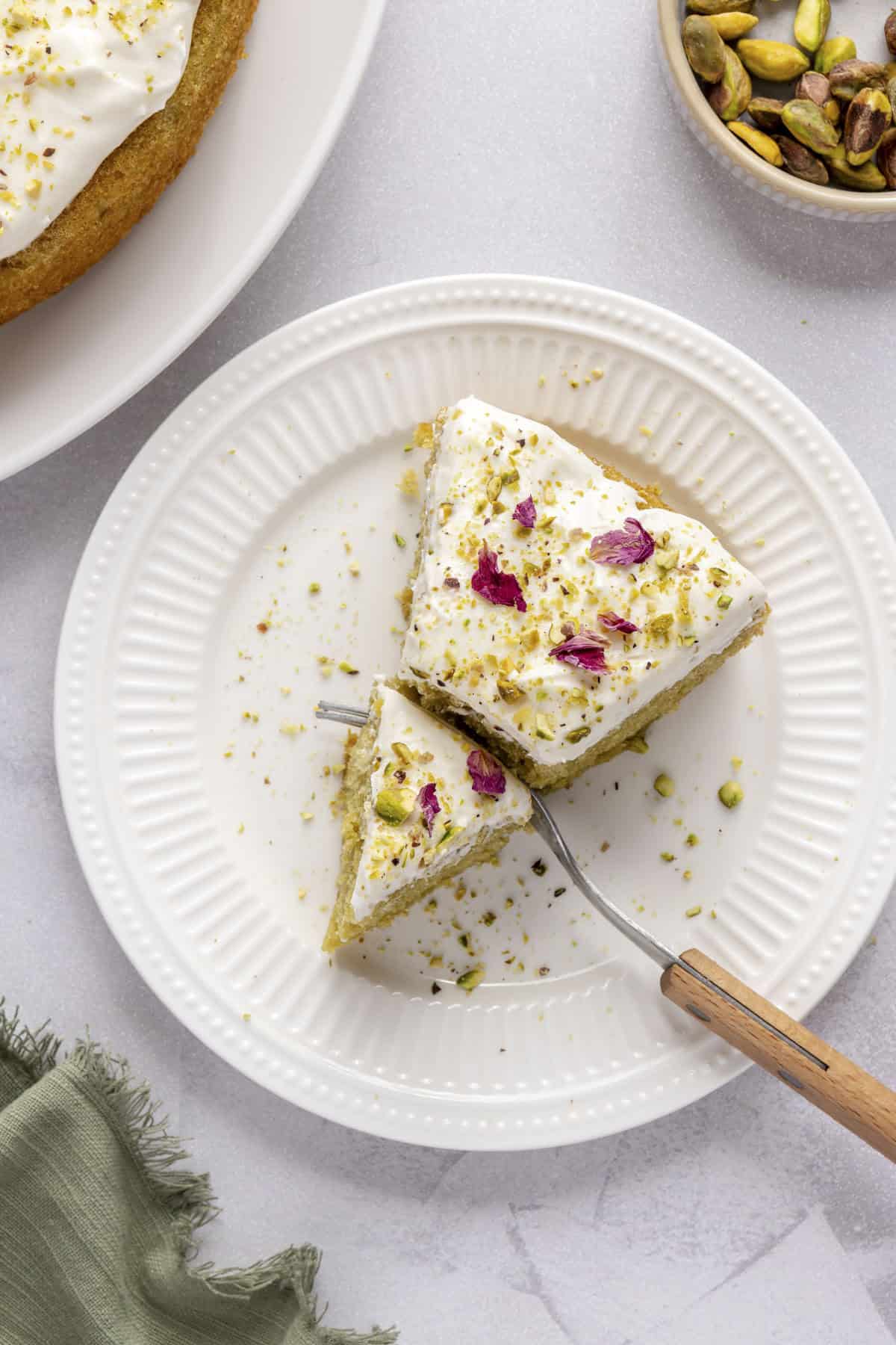 Pistachio cake slice on a small plate with a fork breaking off a piece.