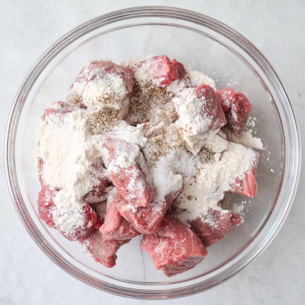 Cubed stew meat in a bowl with flour and seasoning added.