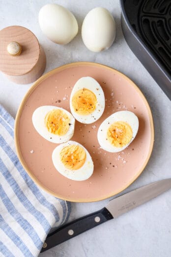 Boiled eggs cut in half on a plate garnished with salt and pepper with air fryer basket nearby.