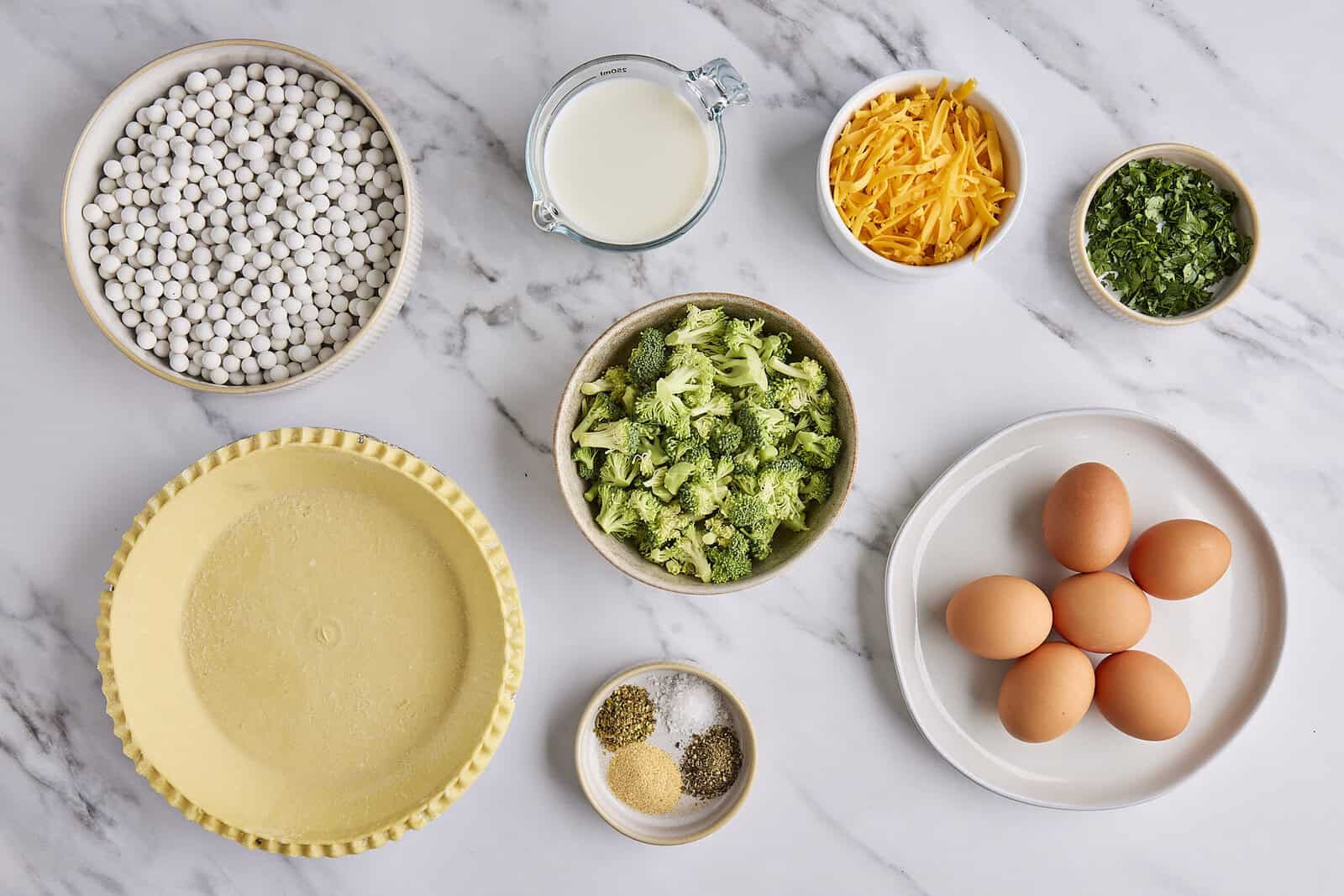 Ingredients for recipe: pie shell, pie weights, milk, broccoli, spices, shredded cheddar cheese, chopped parsley, and eggs.