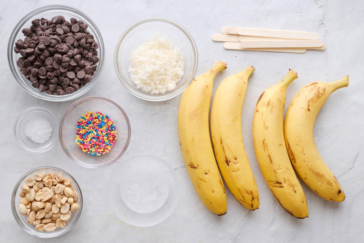 Ingredients for recipe plus optional toppings: chocolate chips, coconut oil, bananas, popsicle skewers, and peanuts, sprinkles shredded coconut, and sea sat.