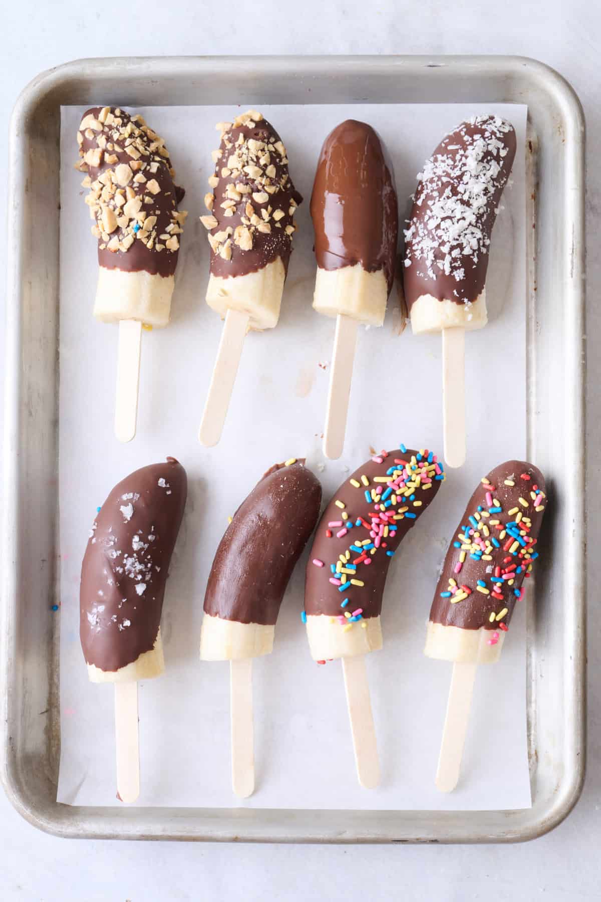 Banana halves dipped in chocolate on parchment paper, only some with toppings, show wet chocolate.