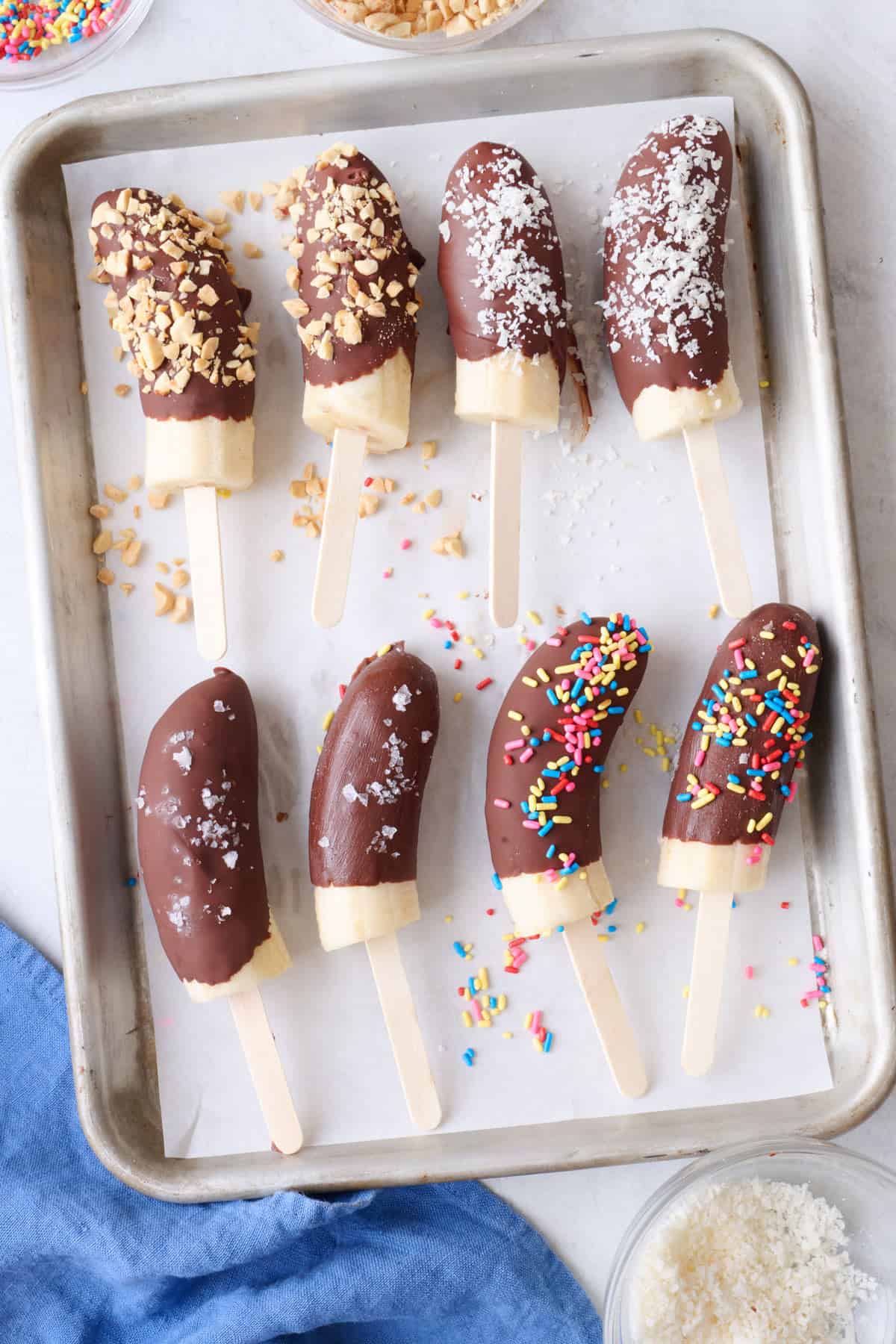 Two of each topping chocolate covered bananas on a clean parchment paper lined sheet pan with extra toppings around: peanut, coconut, sea salt, and sprinkles.