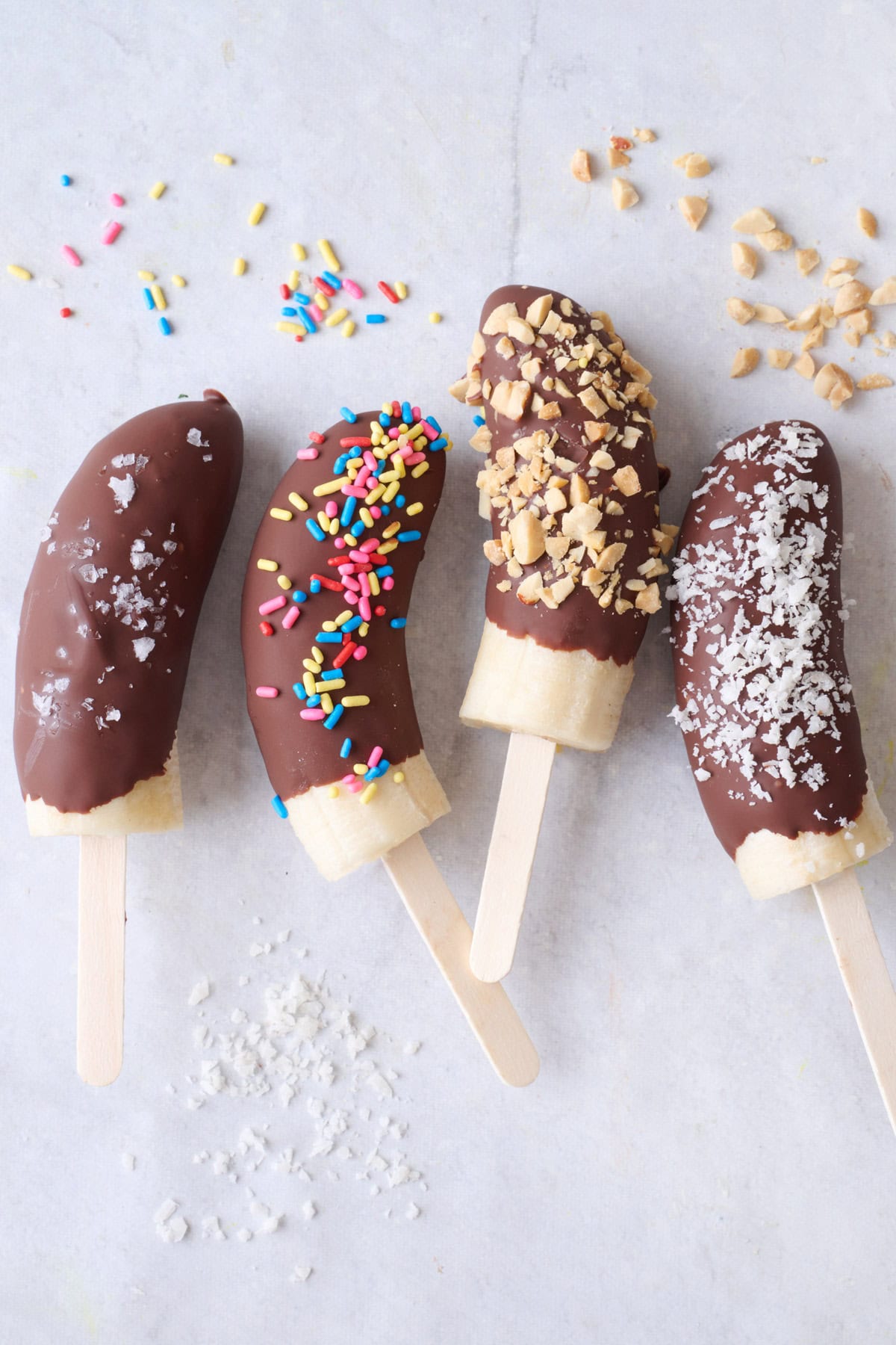 Chocolate covered bananas on a stick with different toppings including sea salt, sprinkles, chopped peanuts and shredded coconut