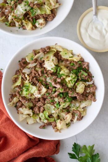 Two shallow bowls of ground beef and cabbage, and yogurt and parsley nearby.