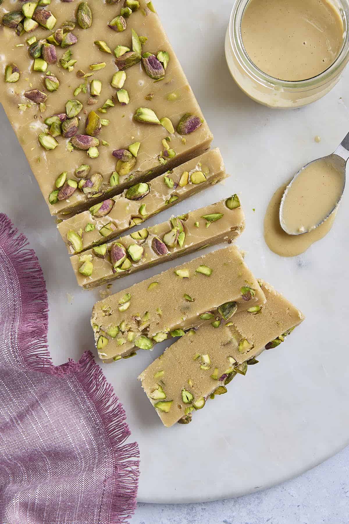 Halva with a few slices cut and a few pieces laying flat to show inside texture, small jar of tahini with small spoon on surface.