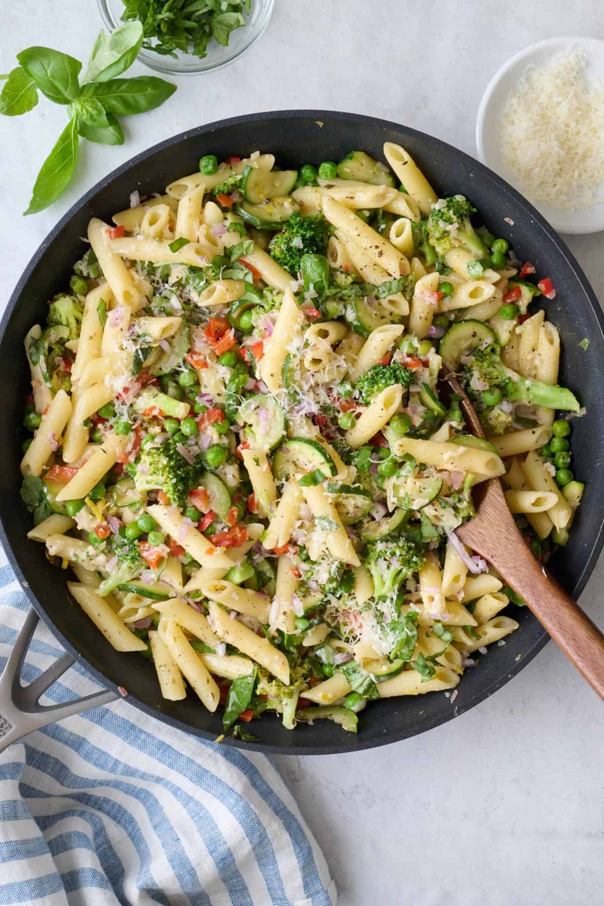 Pasta primavera in skillet garnished with Parmesan cheese and basil, serving spoon dipped inside and small dish of extra parmesan nearby.