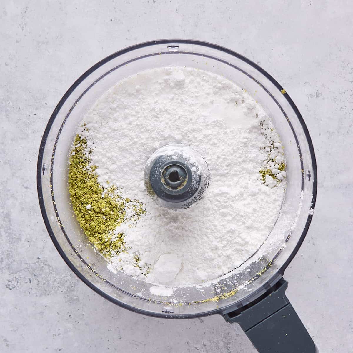 Crushed pistachios in food processor with powdered sugar added on top.