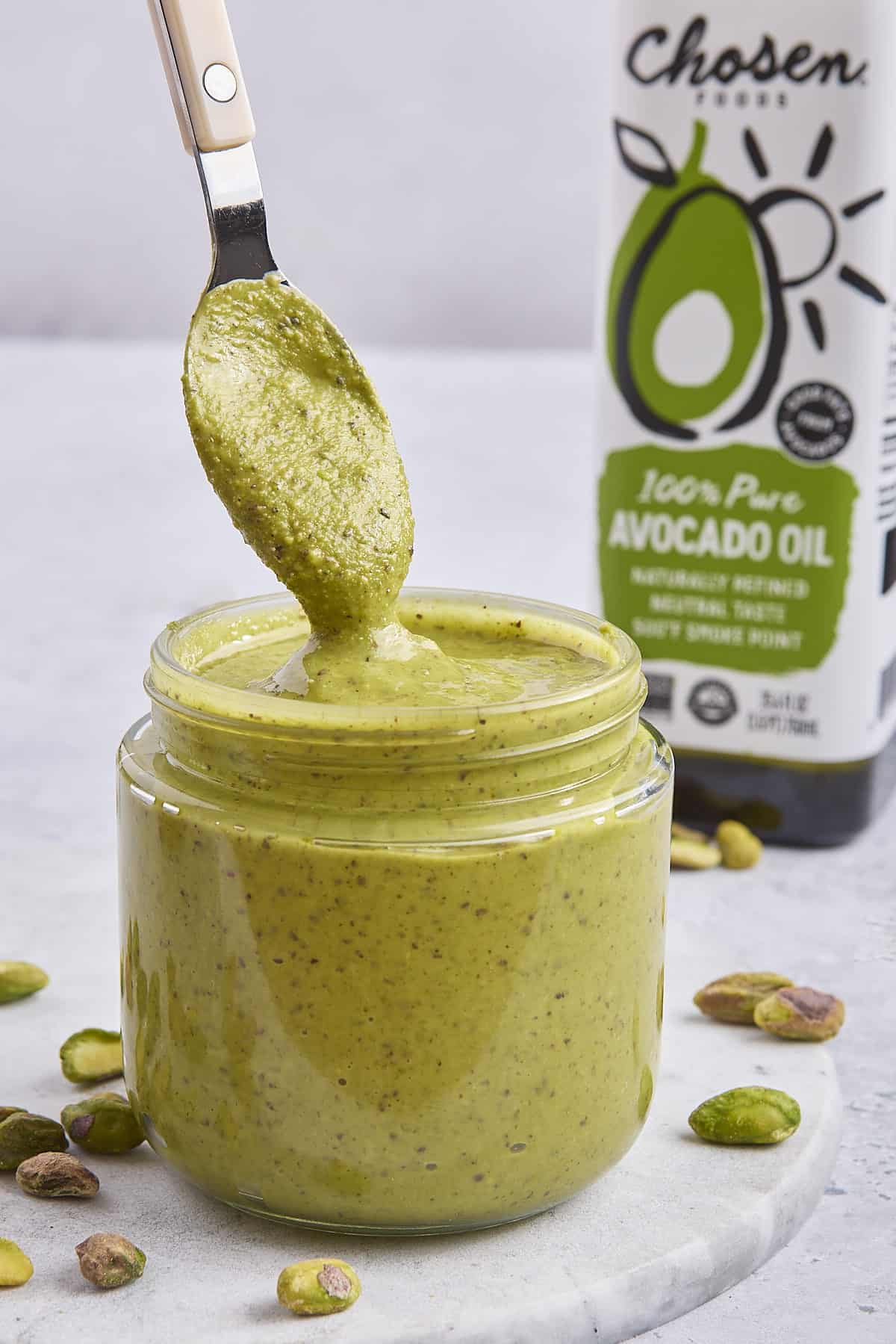 Pistachio cream made with Chosen Foods 100% Pure Avocado Oil in a small jar with a spoon drizzling some back into it.