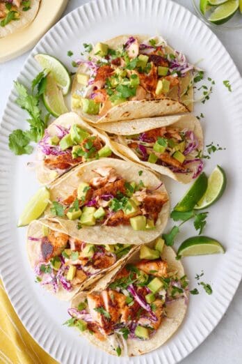 6 assembled salmon tacos with fish taco slaw and diced avocado tight on a long platter with lime wedges.