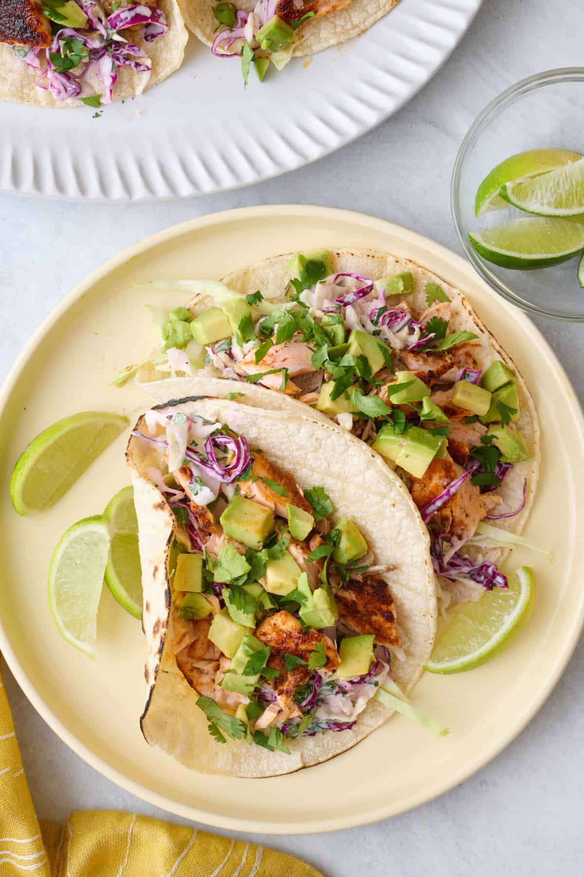 Two assembled, open faced salmon tacos on a small plate with platter nearby.