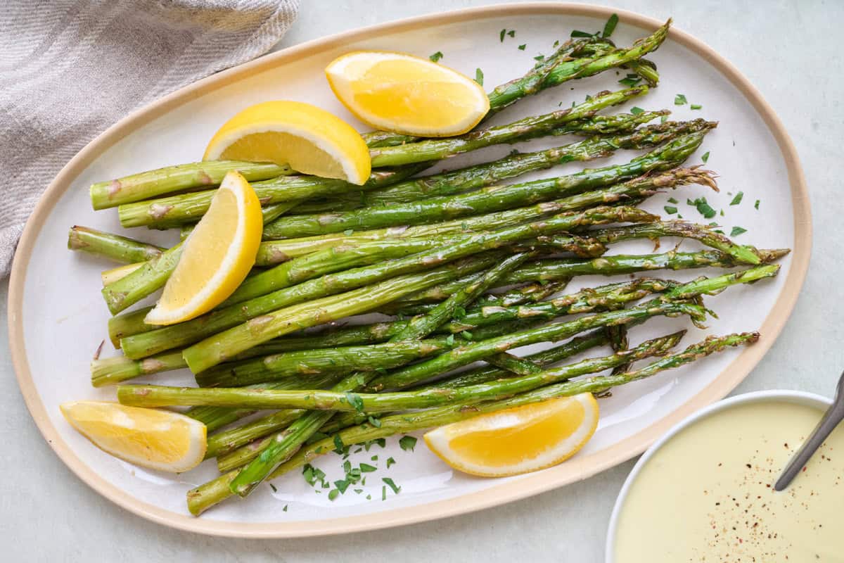 Baked asparagus on a serving dish with a bowl of hollandaise sauce nearby and lemon slices.