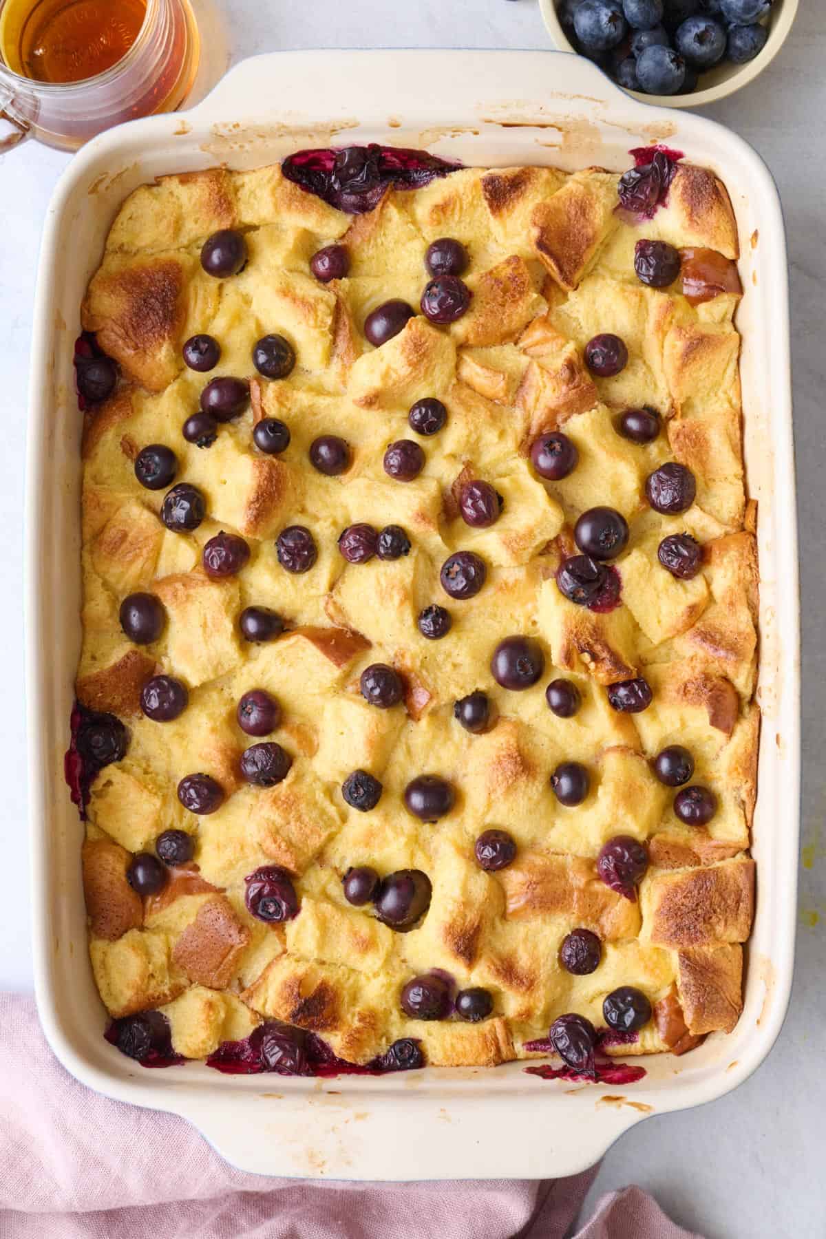 Blueberry french toast casserole in baking dish garnished with dish of syrup, fresh blueberries and plate with fork nearby.