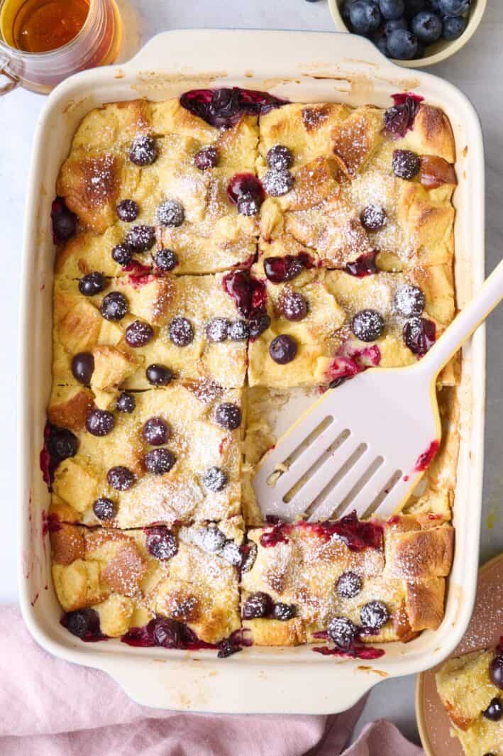 French toast casserole with blueberries dusted with powdered sugar and cut into 8 pieces with a serving removed to a plate on the side, serving spatula/spoon dipped inside.