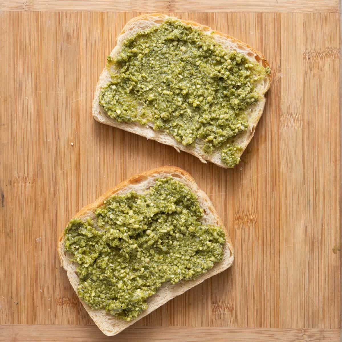 2 bread slices on cutting board with pesto on the pieces.