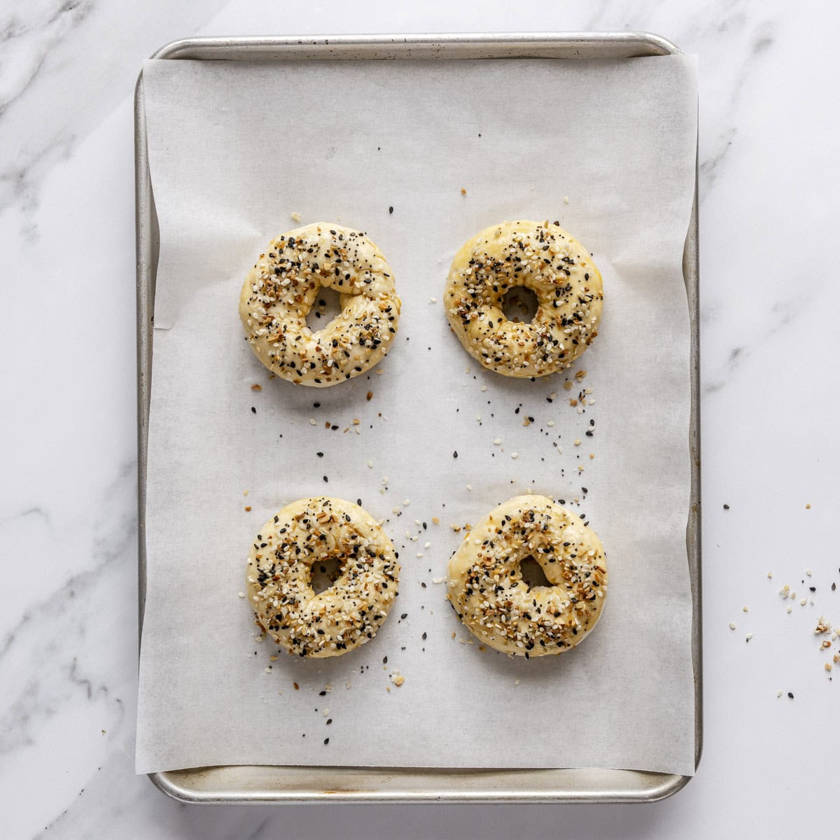 4 bagels on small parchment lined baking sheet with egg wash and everything bagel seasoning before baking.
