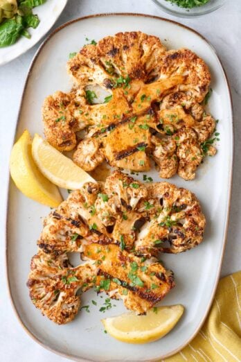 Cauliflower steaks on a small platter with a small dish of chopped parsley, garnished with fresh parsley and lemon wedges.
