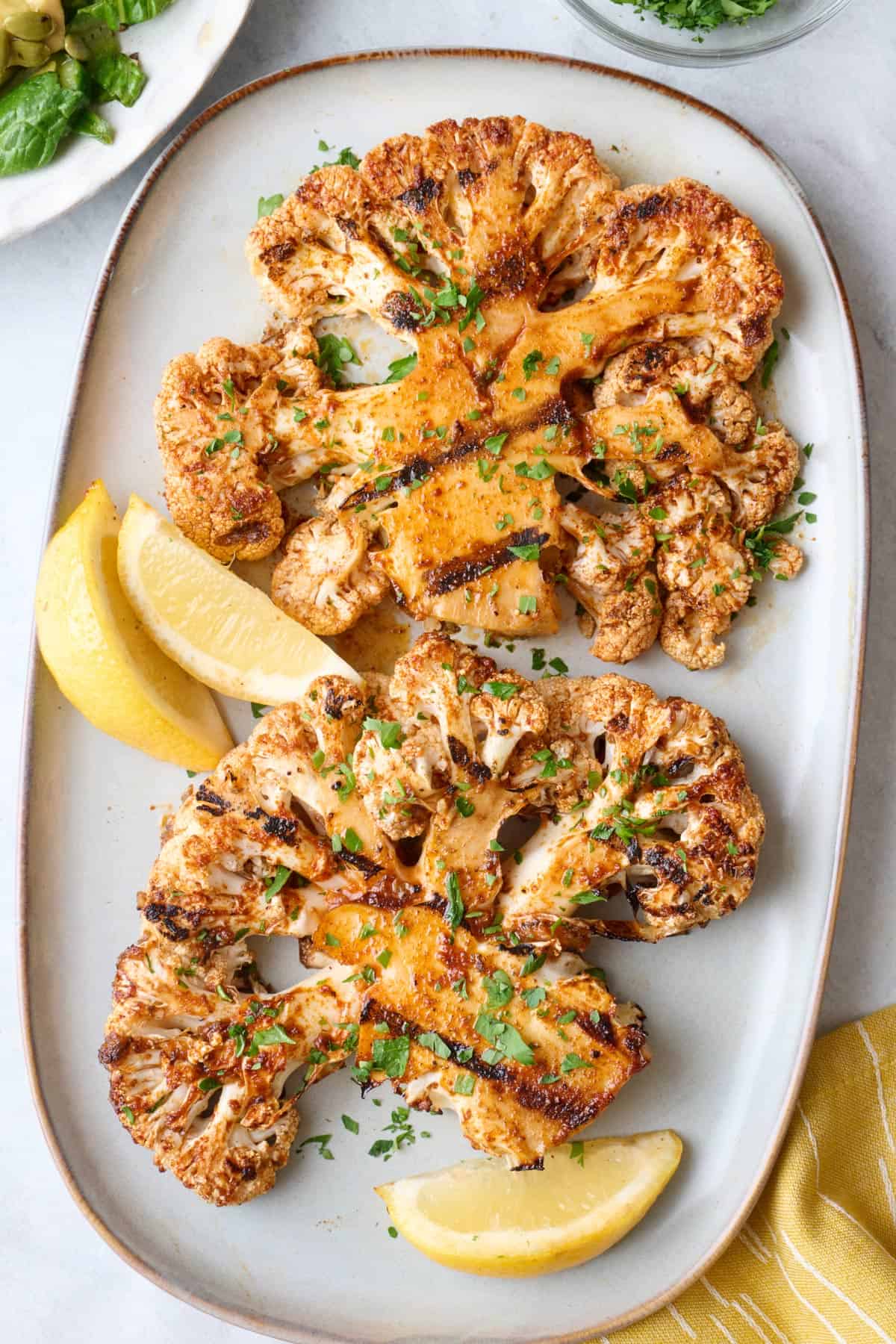 Cauliflower steaks on a small platter with a small dish of chopped parsley, garnished with fresh parsley and lemon wedges.