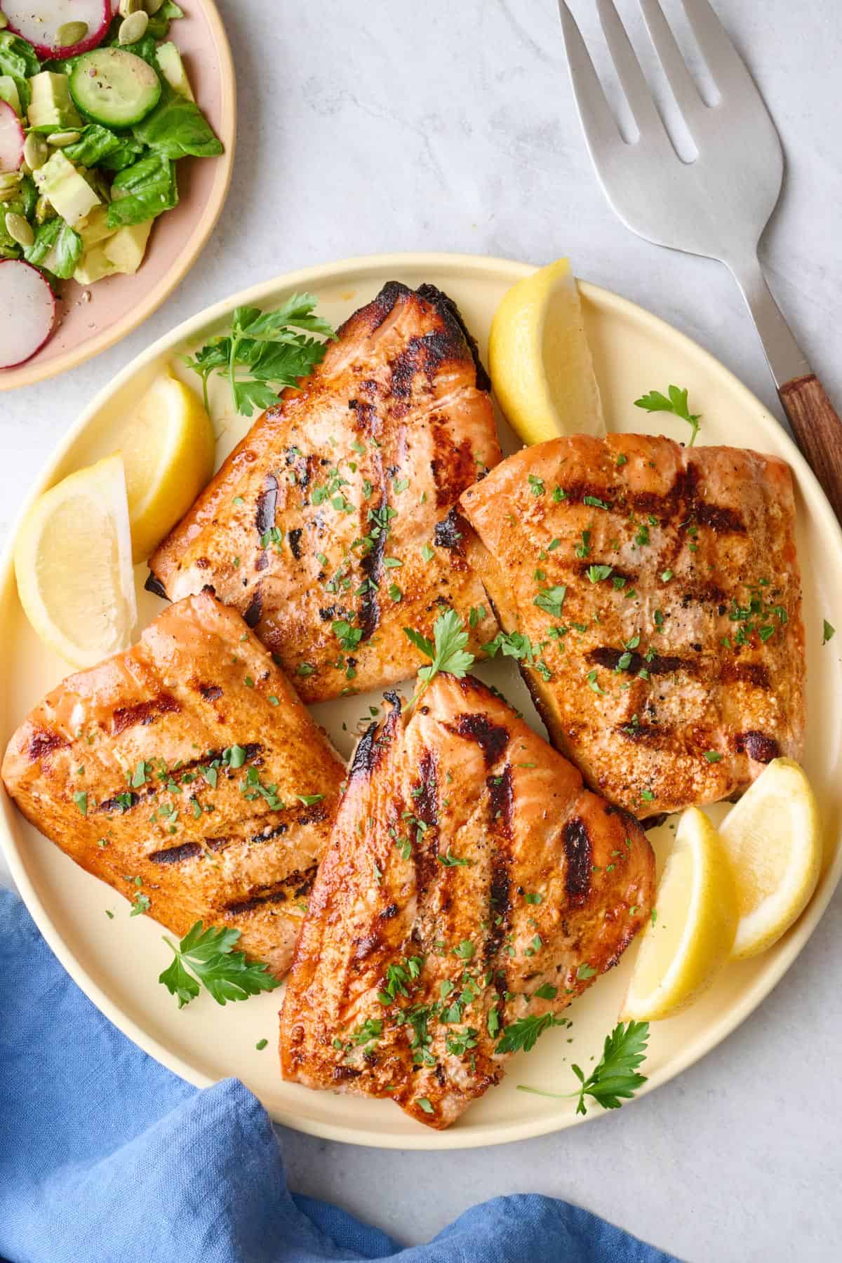 Grilled salmon fillets on a round plate with lemon wedges, garnished with fresh parsley, large serving fork nearby.