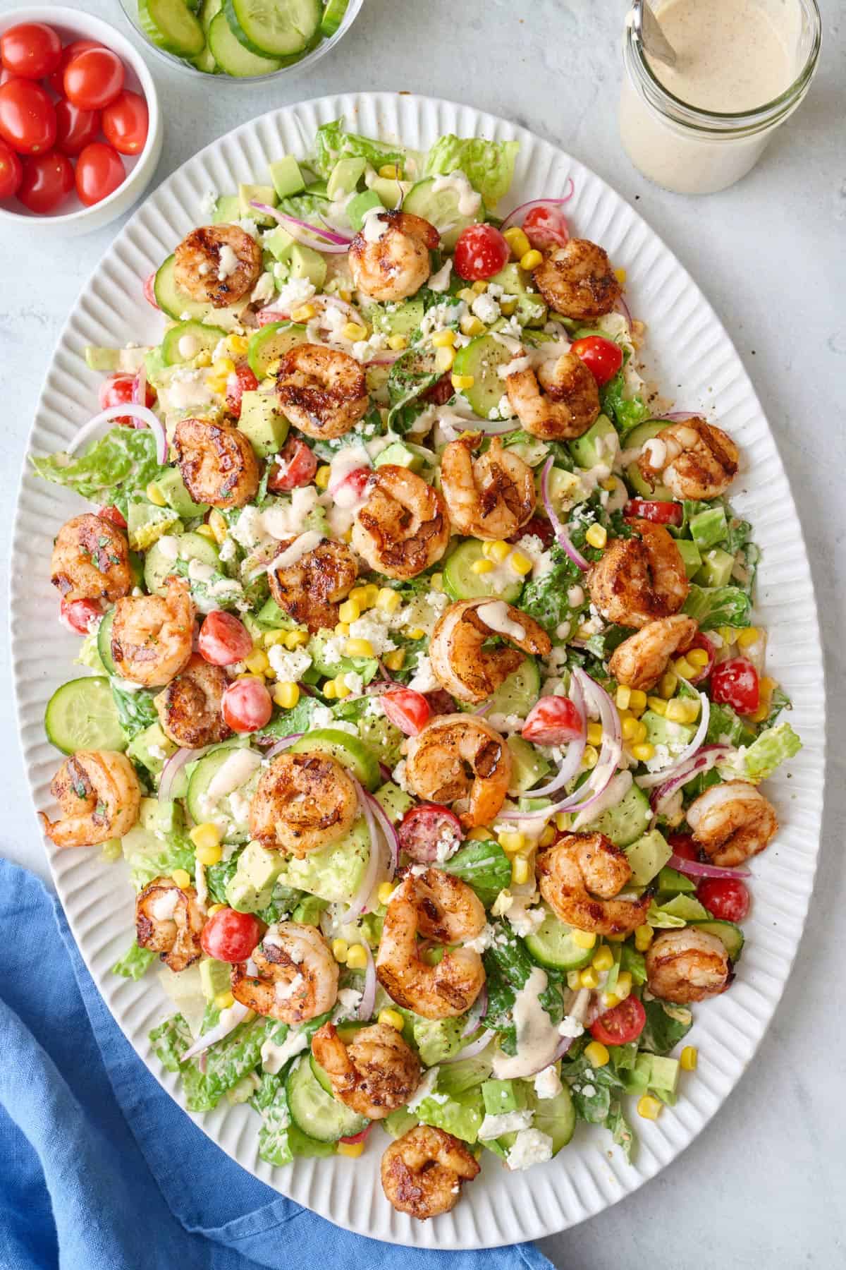 Grilled shrimp salad on platter with extra dressing drizzled on top, salad set dipped into salad, platter at an angle, fresh ingredients around.