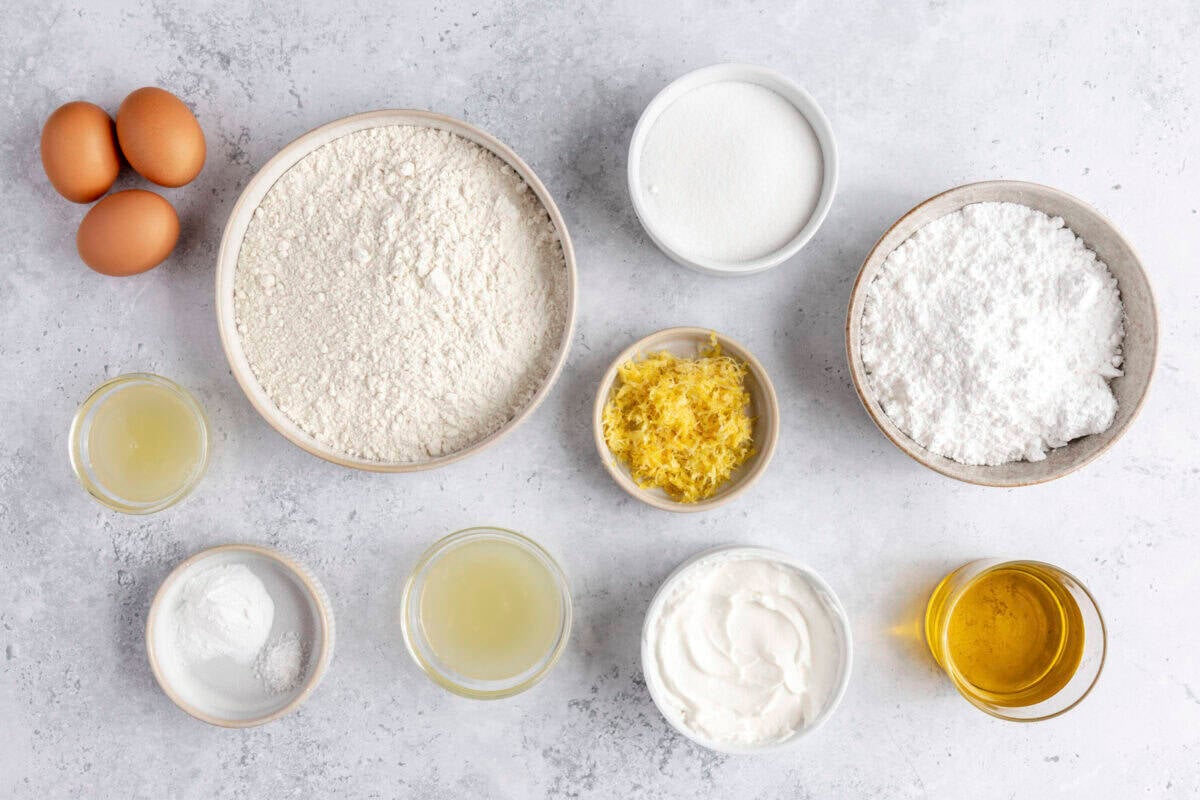 Ingredients for recipe in individual bowls: eggs, flour, sugar, lemon juice, lemon zest, greek yogurt, olive oil, and powdered sugar.