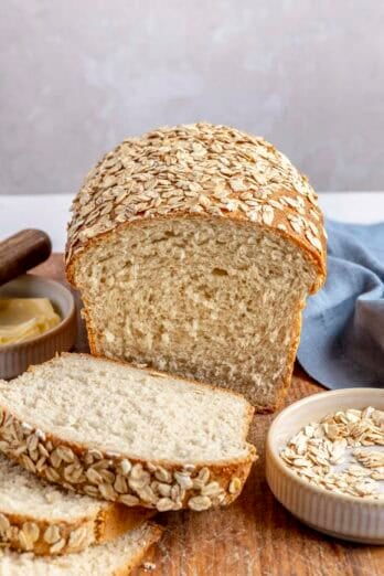 Oatmeal Bread on a cutting board with the top visible, partially sliced with a few slices laying forward, small dish of oats and butter nearby.