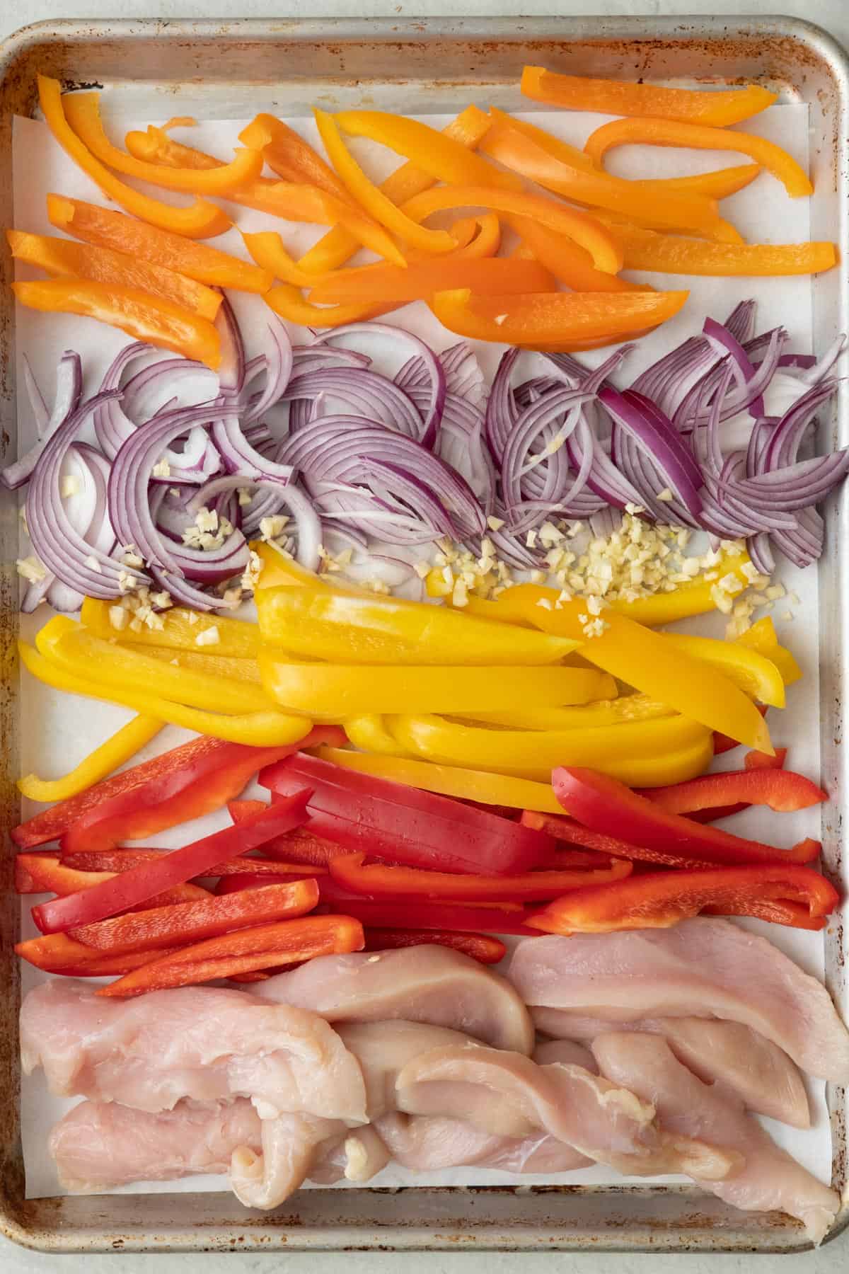 Sliced chicken, peppers, onions, and garlic on a baking sheet (separated into sections).