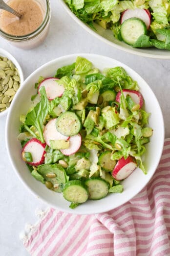 Simple green salad in a serving bowl with a little more dressing drizzled on top, serving bowl and small dish of seeds nearby.