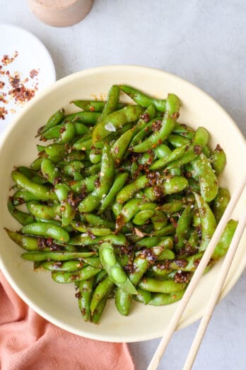 Spicy edamame on plate, a pod opened, with chopsticks resting on plate.