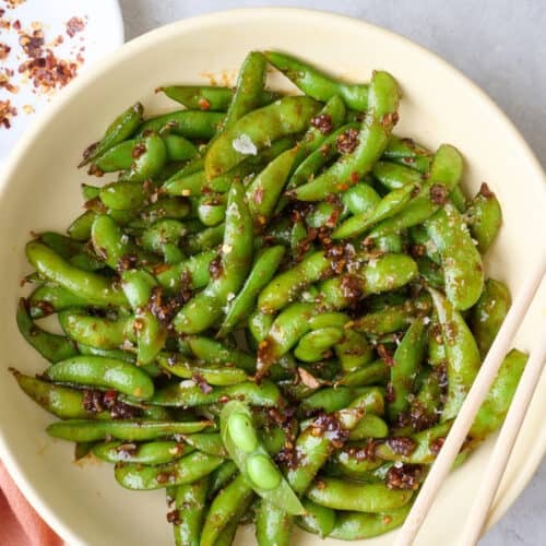 Spicy edamame on plate, a pod opened, with chopsticks resting on plate.