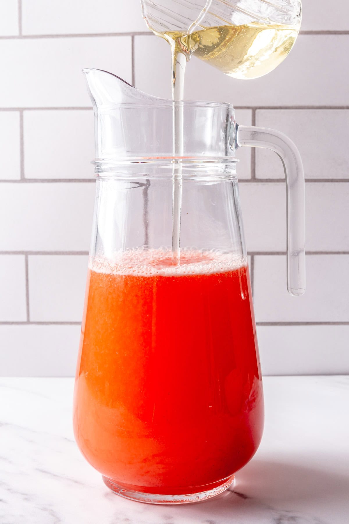 Pouring simple syrup into pitcher of strawberry lemonade.