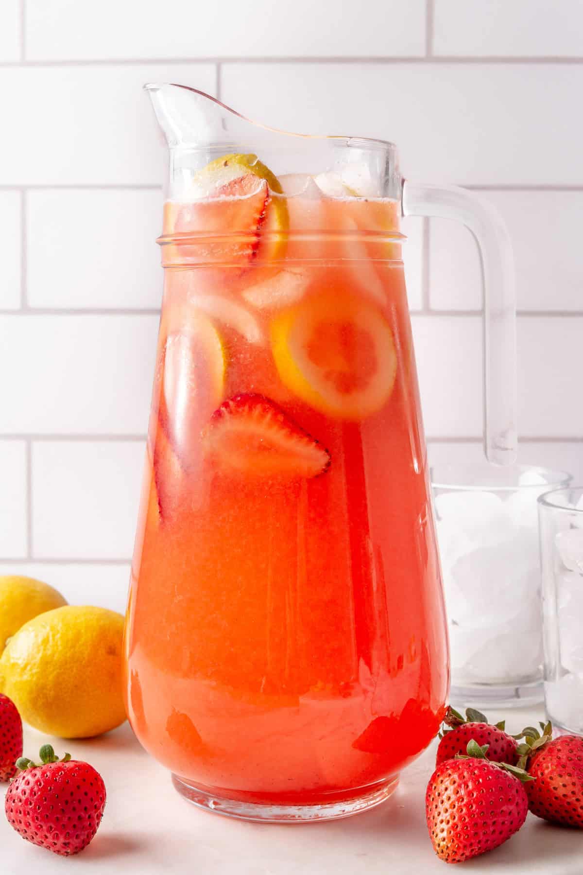 Pitcher of Strawberry lemonade with fresh strawberries, lemons and 1-2 glasses with ice nearby.