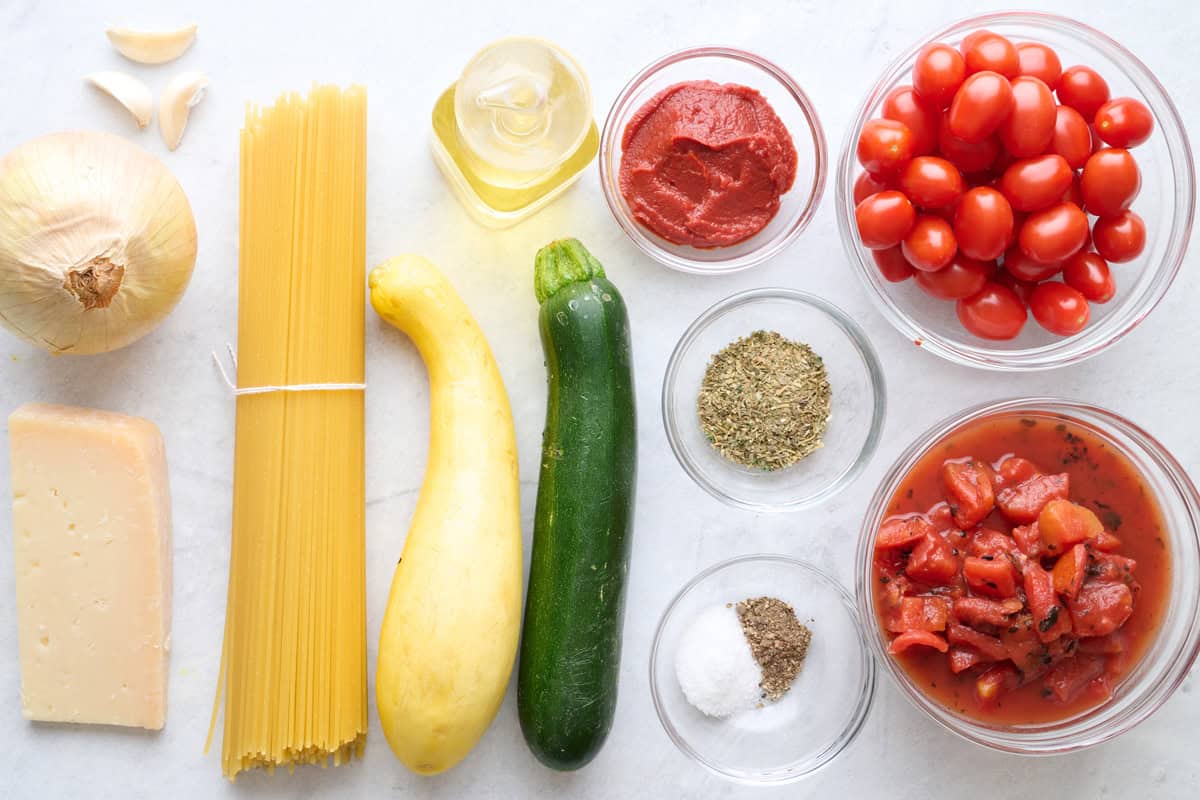 Ingredients for recipe before prepping: garlic, onion, parmesan cheese, uncooked spaghetti, oil, squash, zucchini, oil, tomato paste, spices, cherry tomatoes, and fire roasted tomatoes.