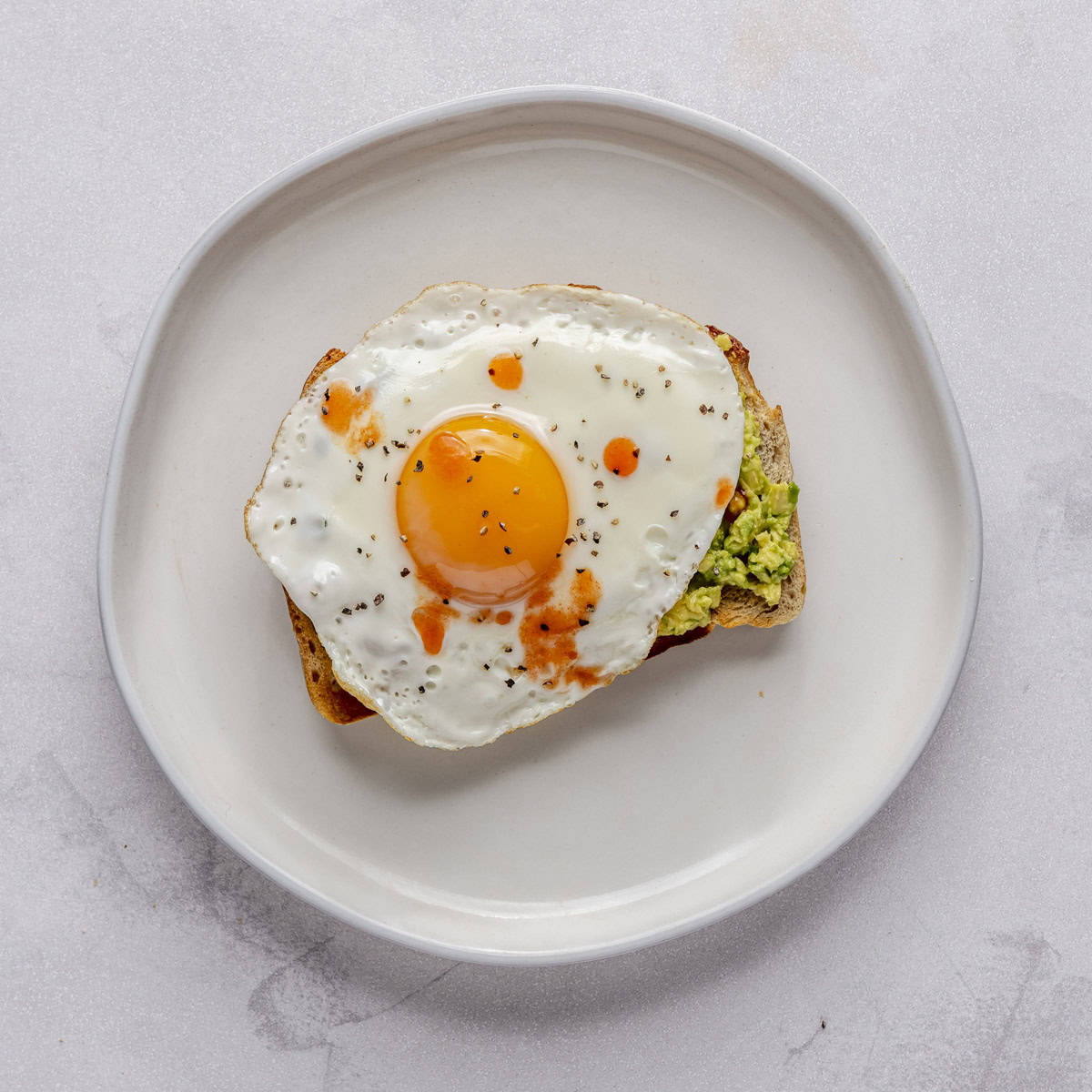 Avocado toast with sunny side up egg with pepper and a few dashes of hot sauce on top.