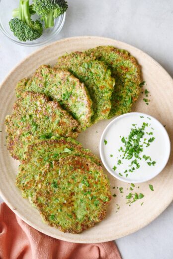 Broccoli fritters on a round plate garnished with a small dish of greek yogurt sauce with chopped parsley on top.
