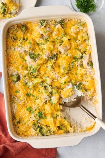 Chicken broccoli rice casserole in a baking dish garnished with chopped parsley, with a serving removed to a plate nearby, and a serving spoon dipped inside casserole dish.