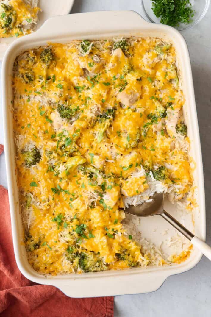 Chicken broccoli rice casserole in a baking dish garnished with chopped parsley, with a serving removed to a plate nearby, and a serving spoon dipped inside casserole dish.