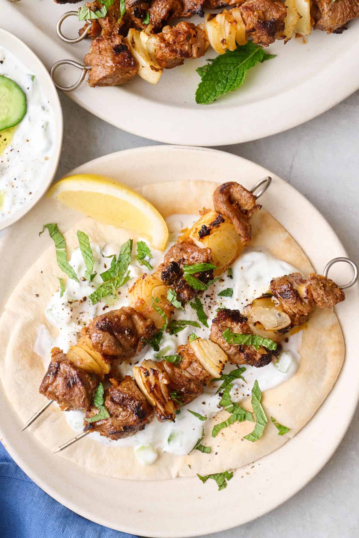 Two lamb skewers served over greek pita sauce smeared on the pita, kabob platter in the background and yogurt bowl on the side.