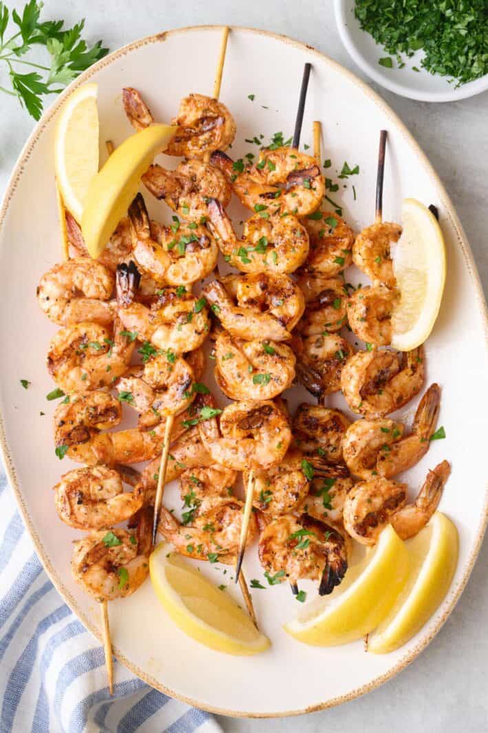 Grilled shrimp skewers on a platter, garnished with chopped parsley and lemon wedges.
