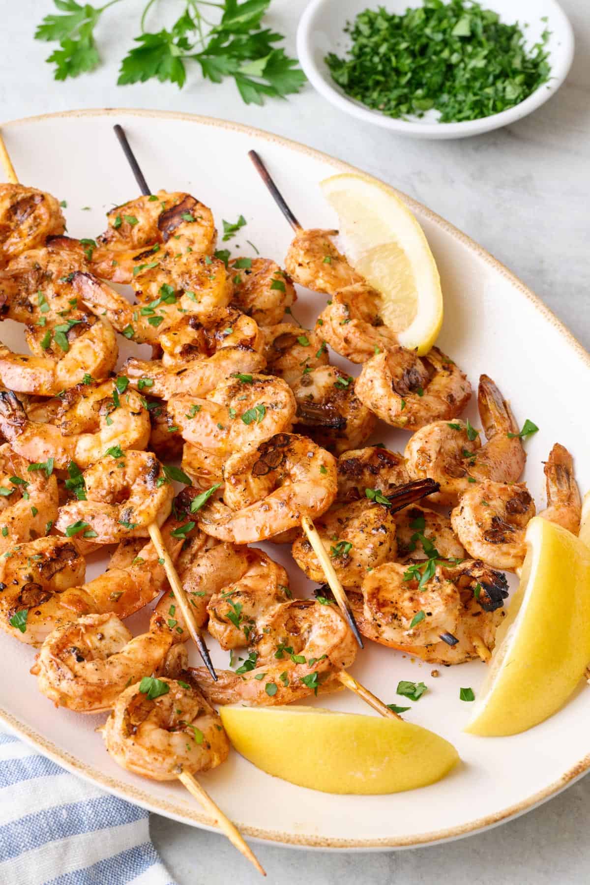 Grilled shrimp skewers on a platter, garnished with chopped parsley and lemon wedges.