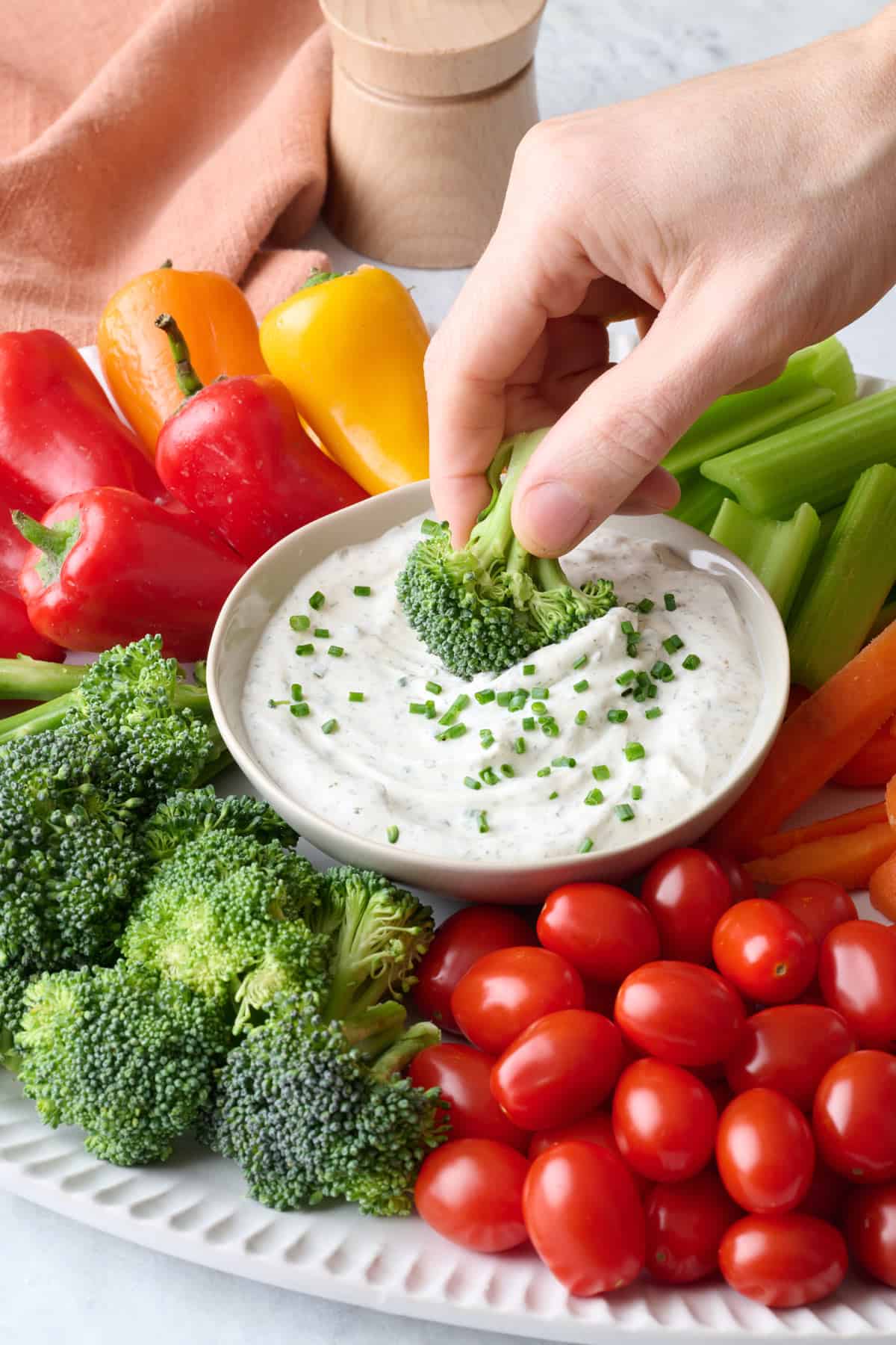 Hand dipping a broccoli florets into a creamy ranch dip on a platter with other vegetables.