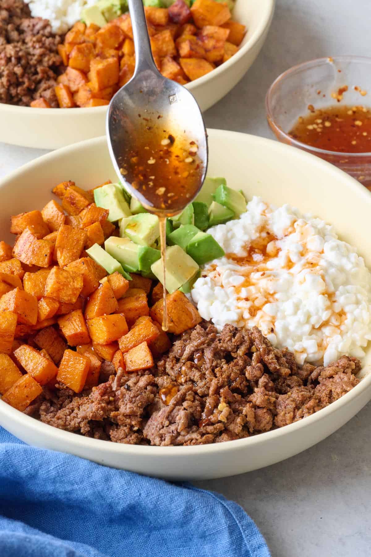 Hot honey being drizzled on a ground beef bowl with cottage cheese, roasted sweet potatoes and diced avocado.