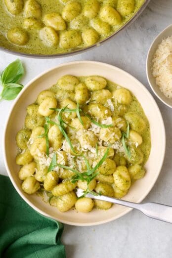 Serving of easy pesto gnocchi in a bowl, garnished with basil and parmesan with skillet of gnocchi nearby.