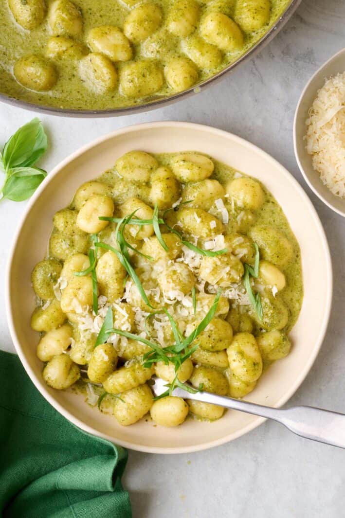 Serving of easy pesto gnocchi in a bowl, garnished with basil and parmesan with skillet of gnocchi nearby.