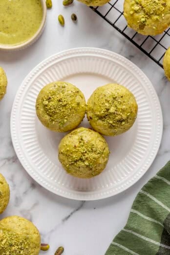 Pistachio muffins topped with glaze and crushed pistachios on a plate with more muffins and glaze nearby.